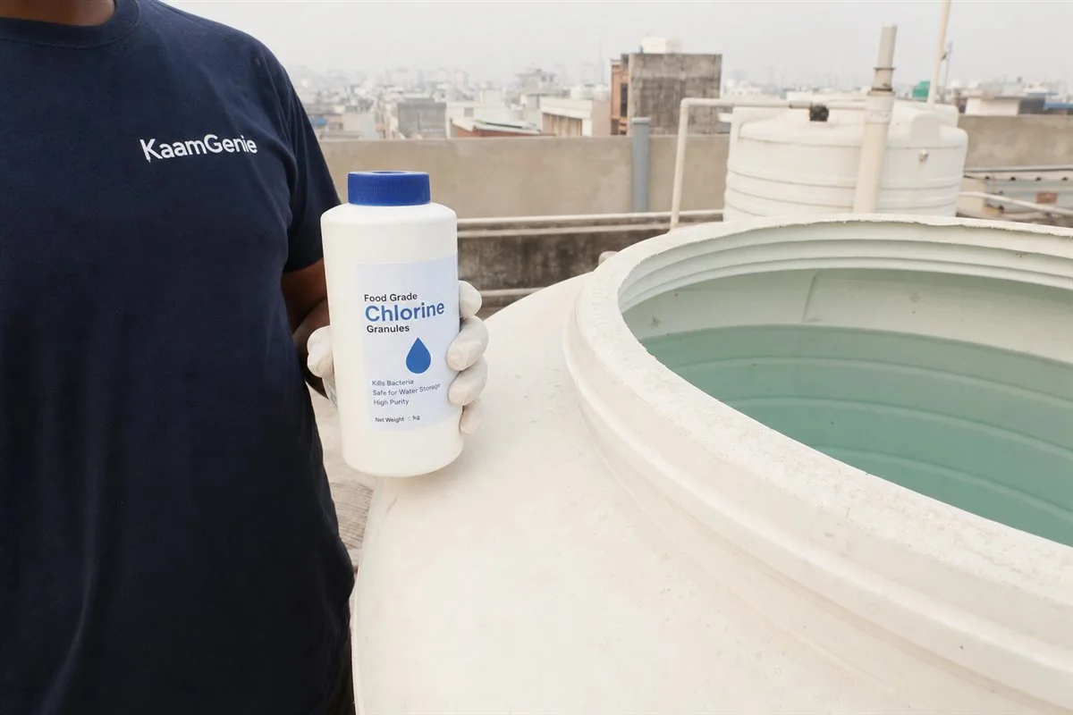 A gloved hand in a navy KaamGenie shirt holding a bottle of food-grade chlorine beside a freshly cleaned Noida water tank