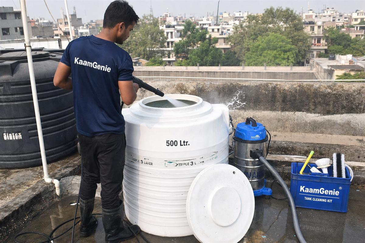 KaamGenie technician in a navy-blue crew shirt cleaning a small 500-litre plastic overhead water tank on a Delhi DDA flat rooftop