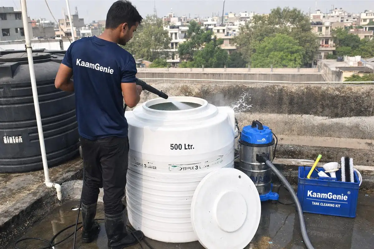 KaamGenie technician in a navy-blue crew shirt cleaning a small 500-litre plastic overhead water tank on a Delhi DDA flat rooftop