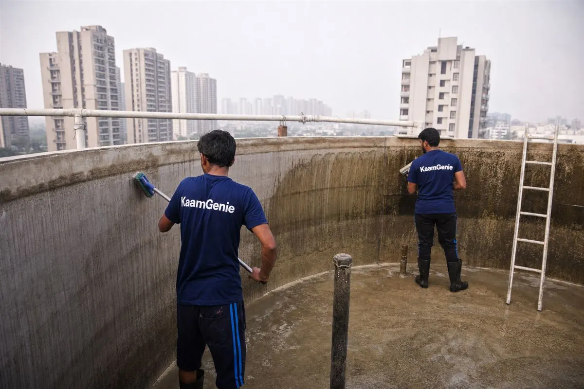 Two KaamGenie workers in navy shirts scrubbing calcium scale and rust stains from a society tower rooftop tank in Greater Noida