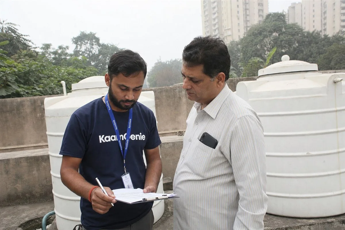 KaamGenie worker in a navy shirt and an AOA member reviewing a tank cleaning compliance record beside a Noida society water tank