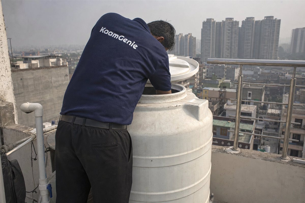 A KaamGenie worker in a navy shirt cleaning the rooftop tank that supplies the kitchen of a Noida home
