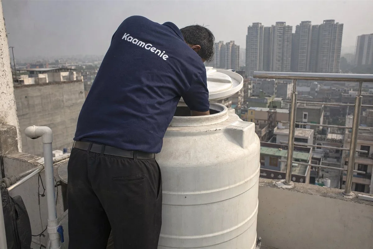 A KaamGenie worker in a navy shirt cleaning the rooftop tank that supplies the kitchen of a Noida home