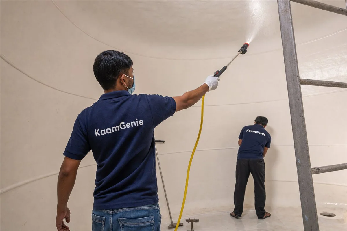 A worker in a navy KaamGenie shirt spraying disinfectant across the clean interior walls of a rooftop water tank on a Noida building