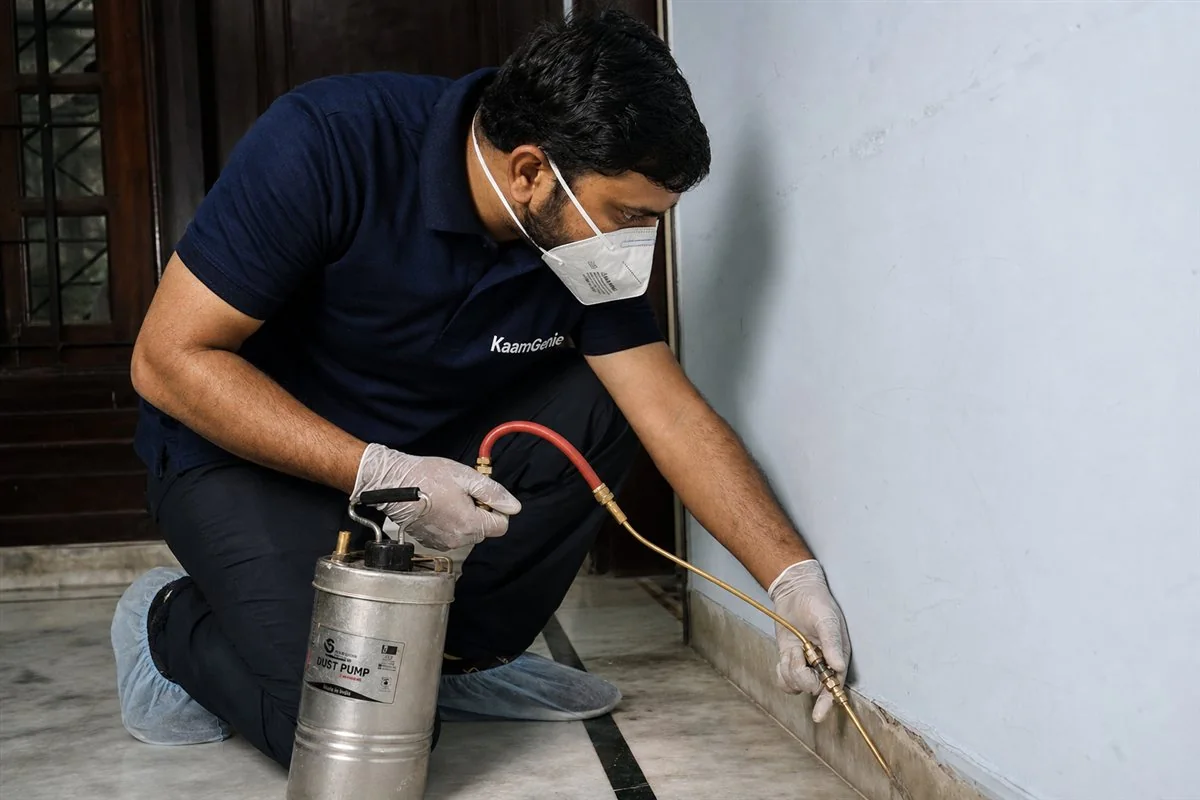 A KaamGenie technician opening lower kitchen cabinets and checking the gap behind the gas stove with a torch during a Delhi pest control inspection
