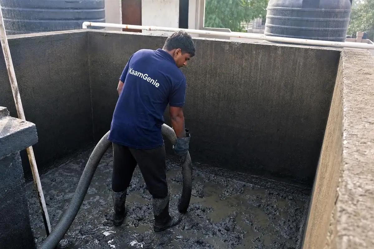 A KaamGenie worker in a navy shirt vacuuming dark sludge from a drained rooftop tank in Noida